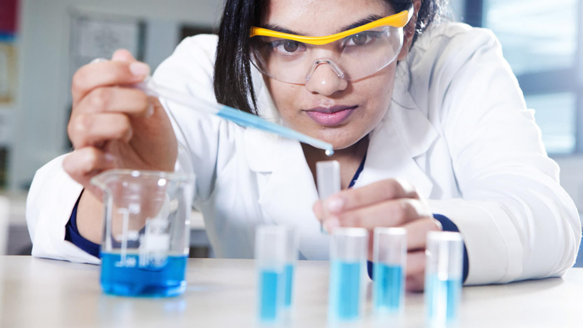 Young woman wearing safety goggles and a white lab coat, holding a dripper and adding the blue liquid to a test tube. There is a beaker with blue liquid on the desk also.