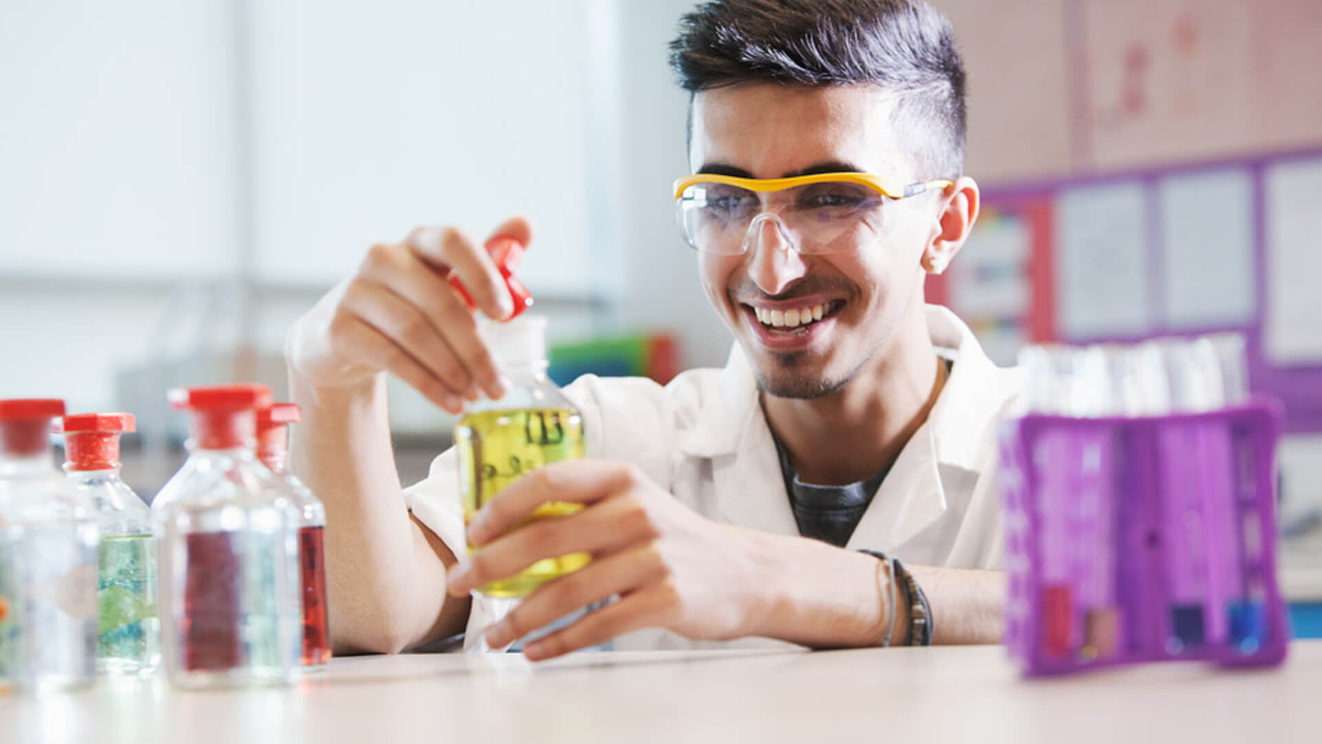 Young man wearing a white lab coat and safety goggles holding a bottle of coloured liquid, doing a science experiment.