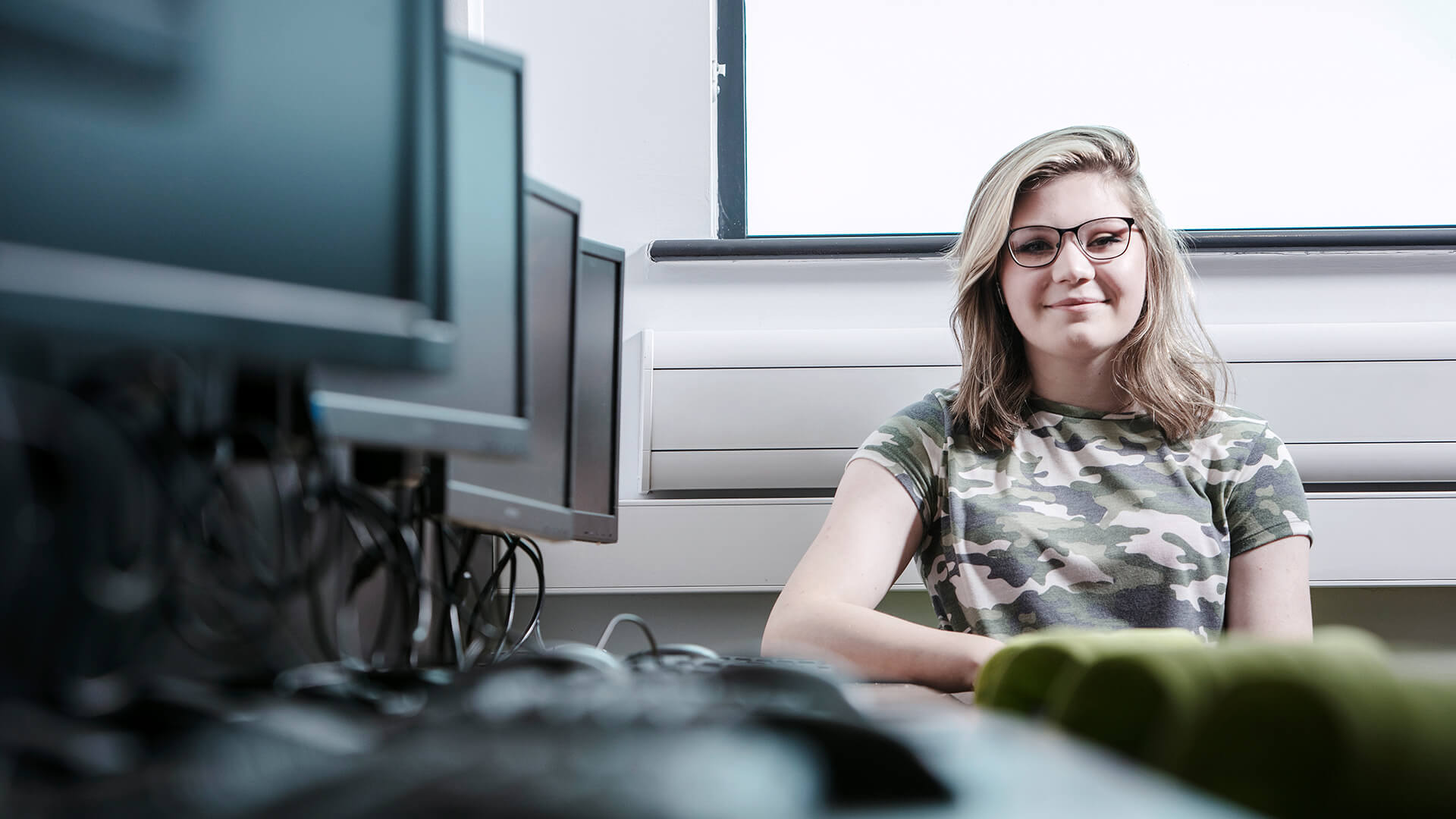 Young woman smiling and sat next to a row of computer screens.