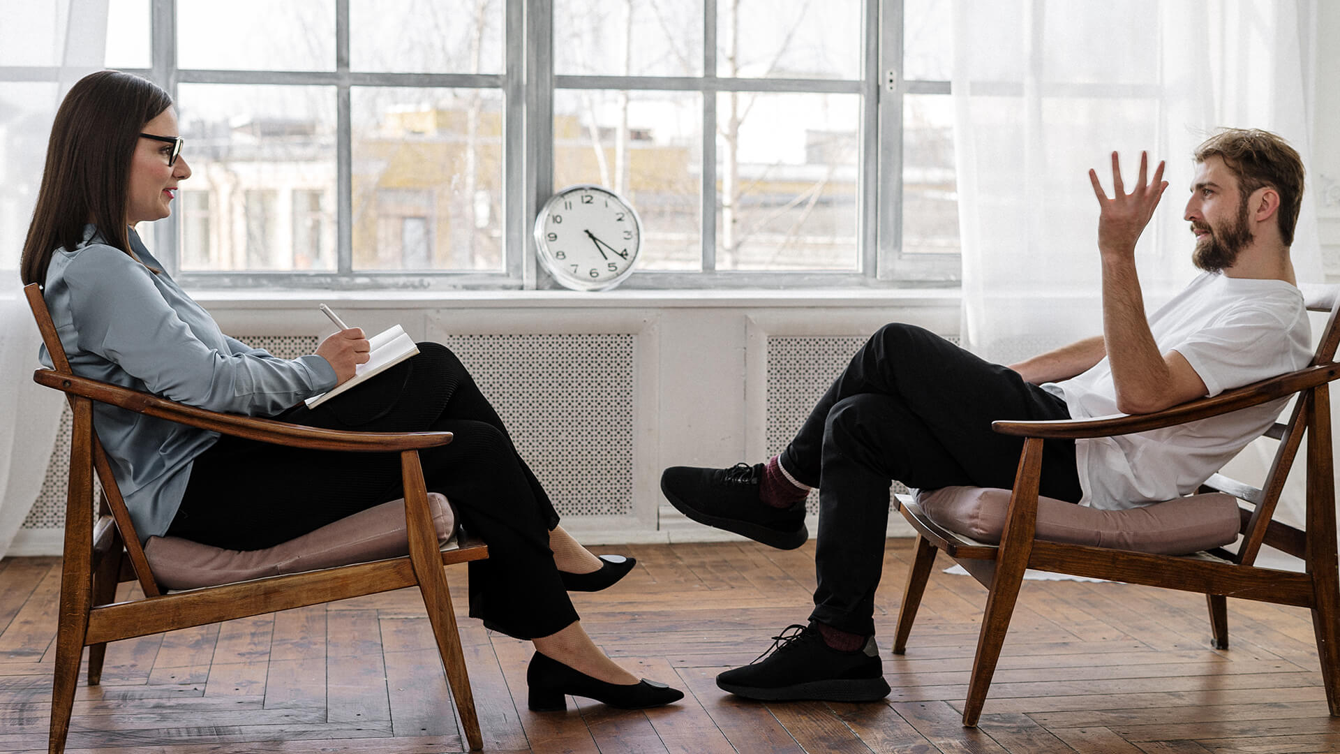 A woman and man sat in chairs opposite to each other having a conversation. The woman is holding a notepad and pen, taking notes.