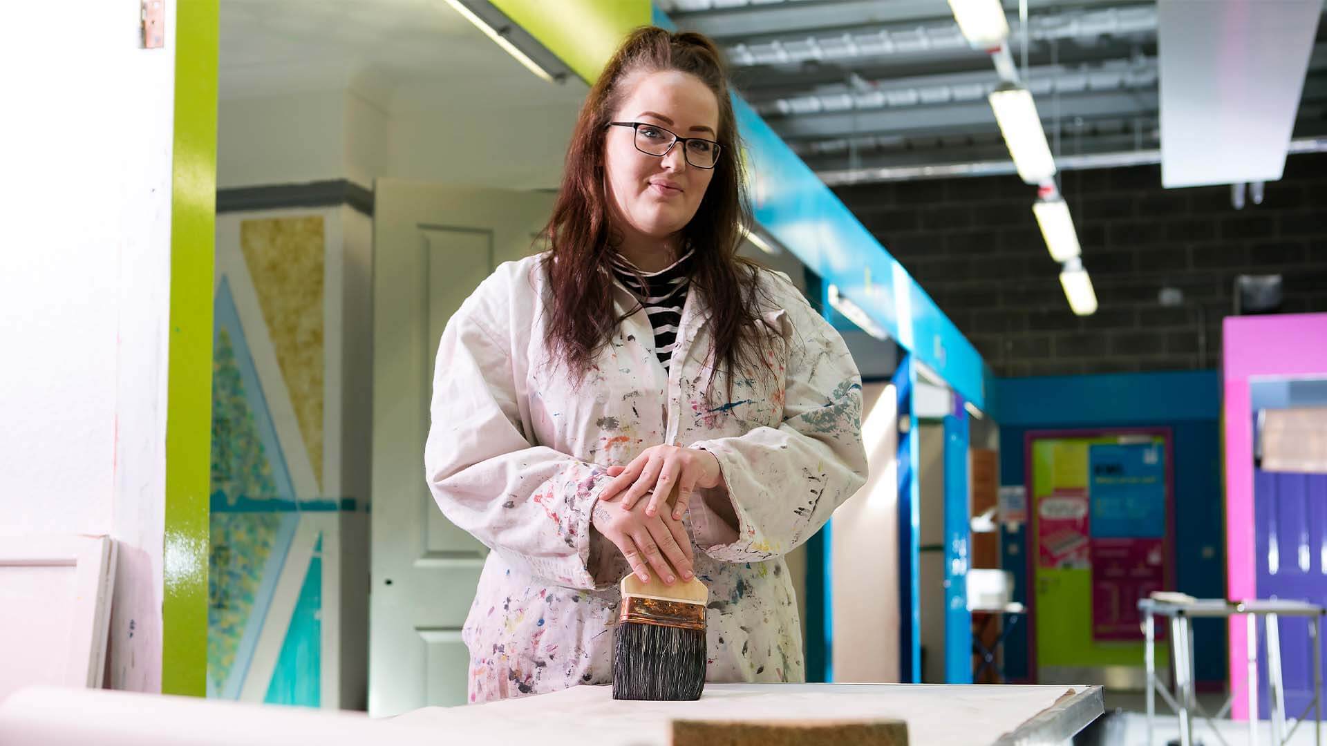 Woman wearing white overalls that are covered in paint splatters, holding a paint brush and smiling.