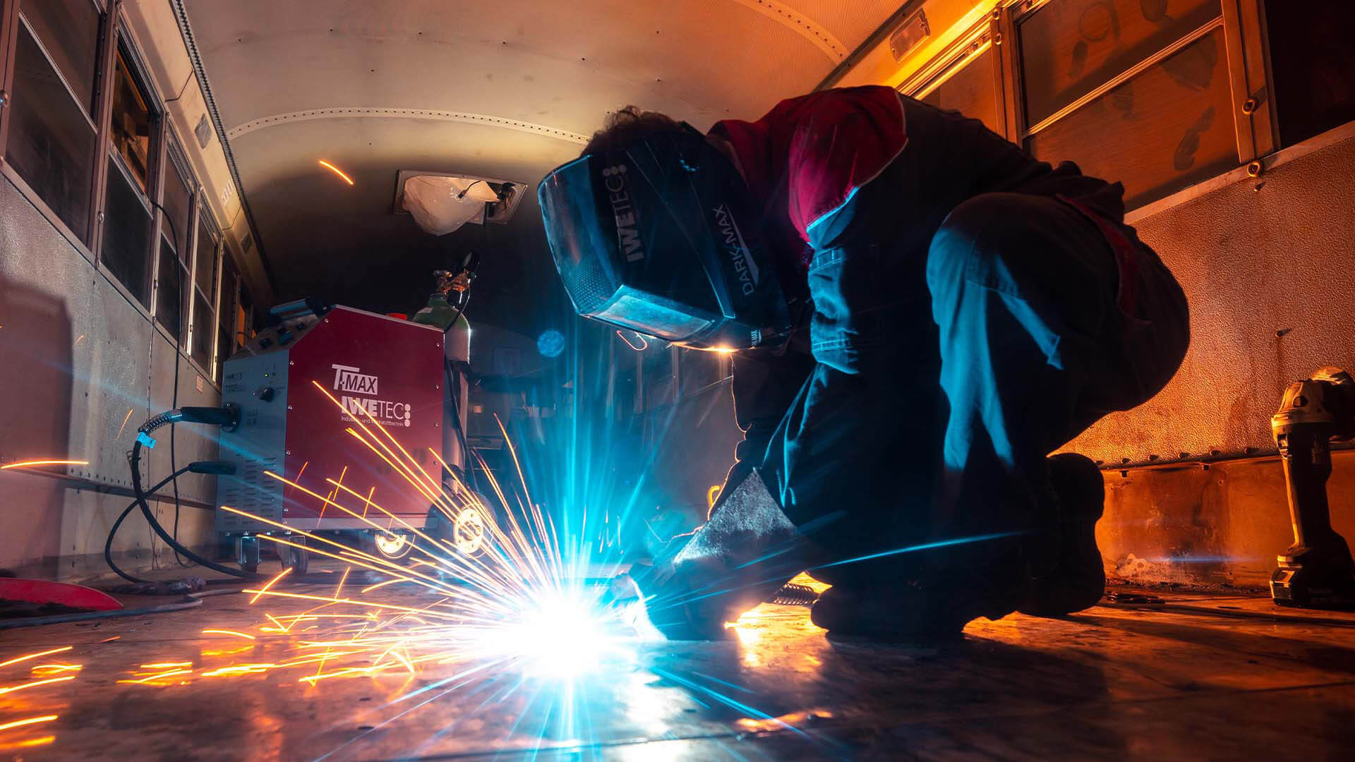 A person wearing a welding helmet and safety overalls, welding a piece of equipment.