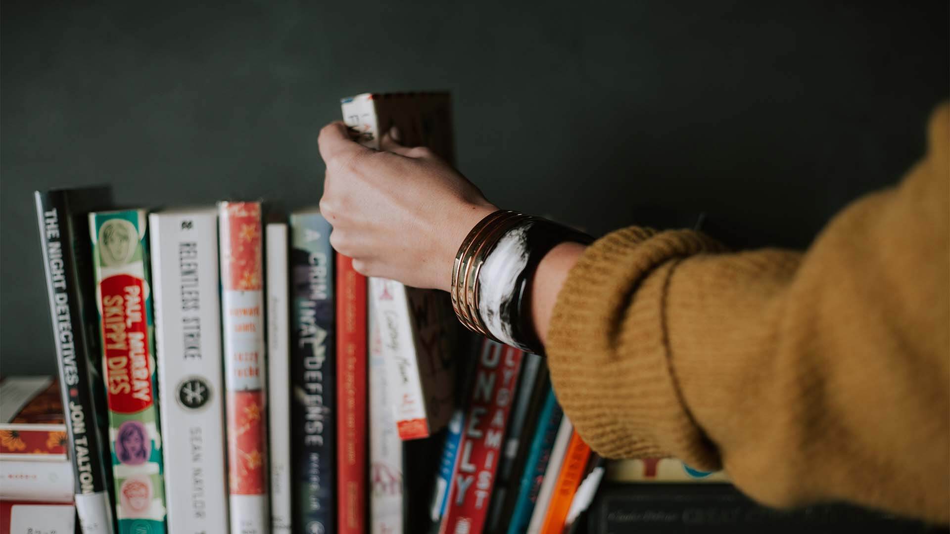 A womans hand grabbing a book from a row of various books on a shelf.