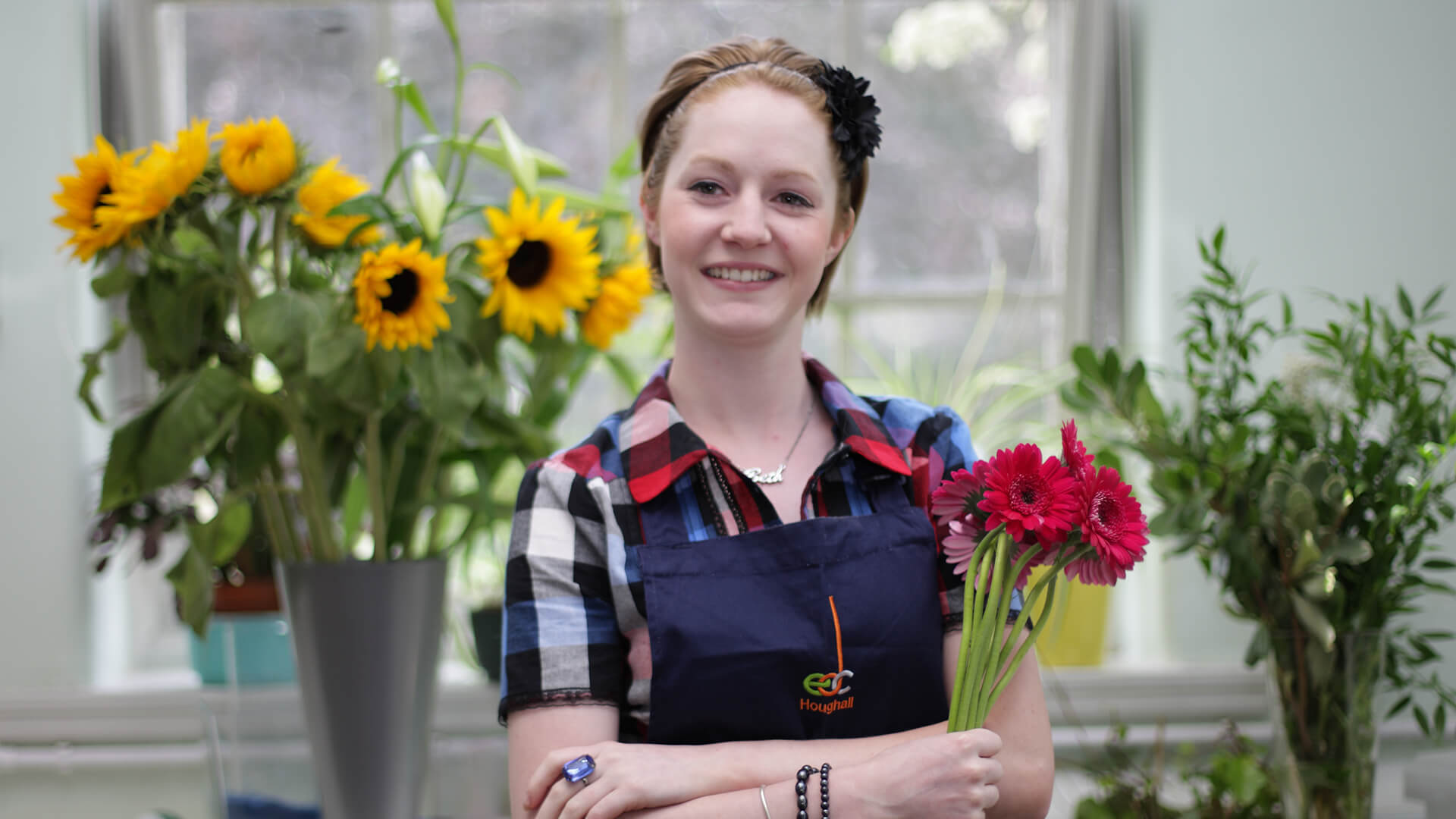 Woman holding a bunch of pink flowers and smiling. There is a bouqet of sunflowers behind her.