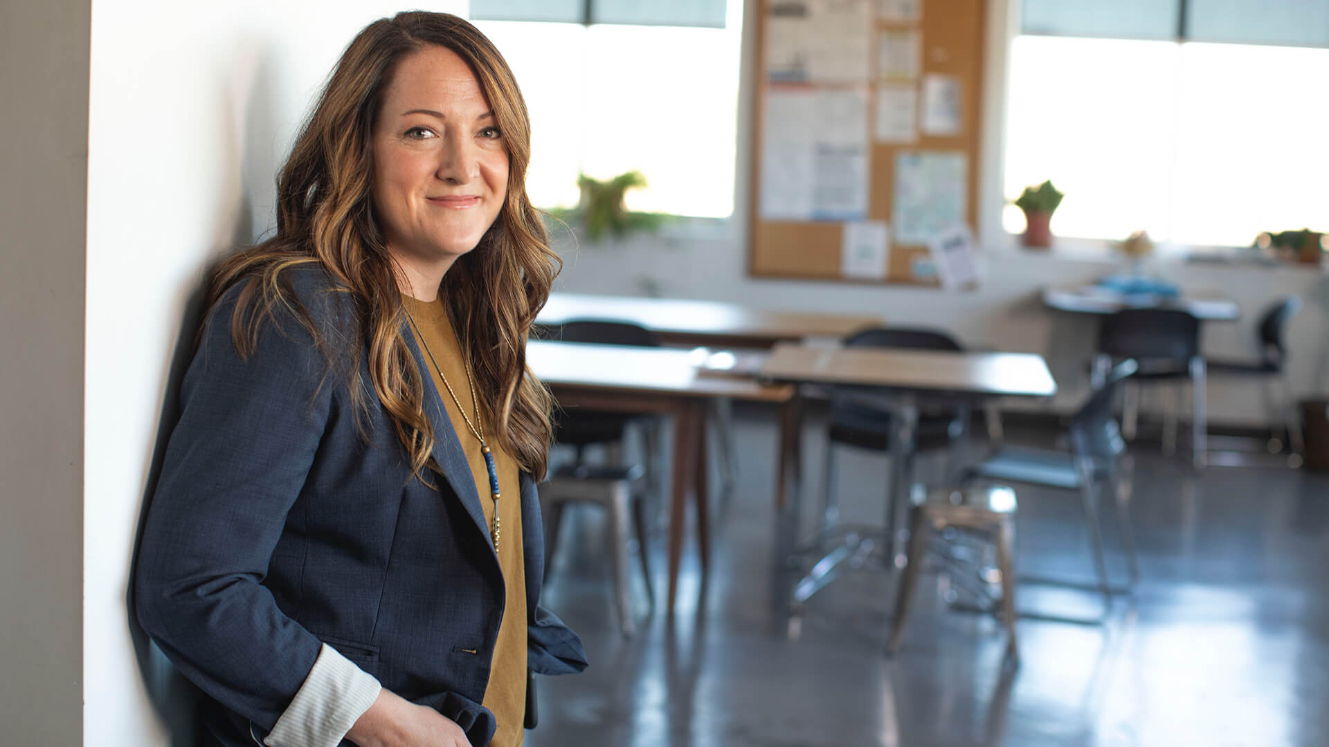 A woman in smart clothing leaning against a wall and smiling with classroom desks behind her.