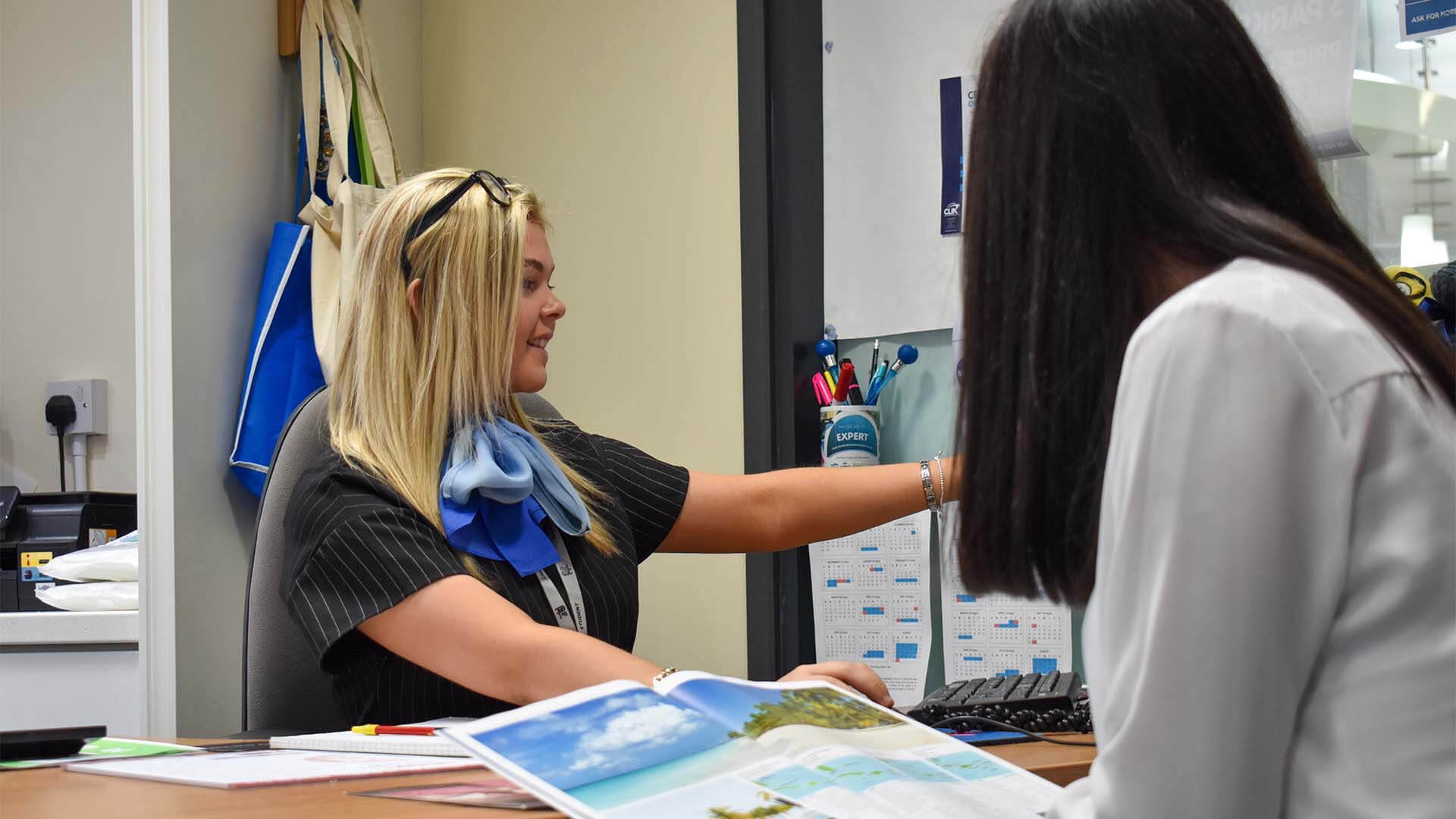 Student working in a travel shop, showing a customer a holiday deal on the computer in a travel shop.