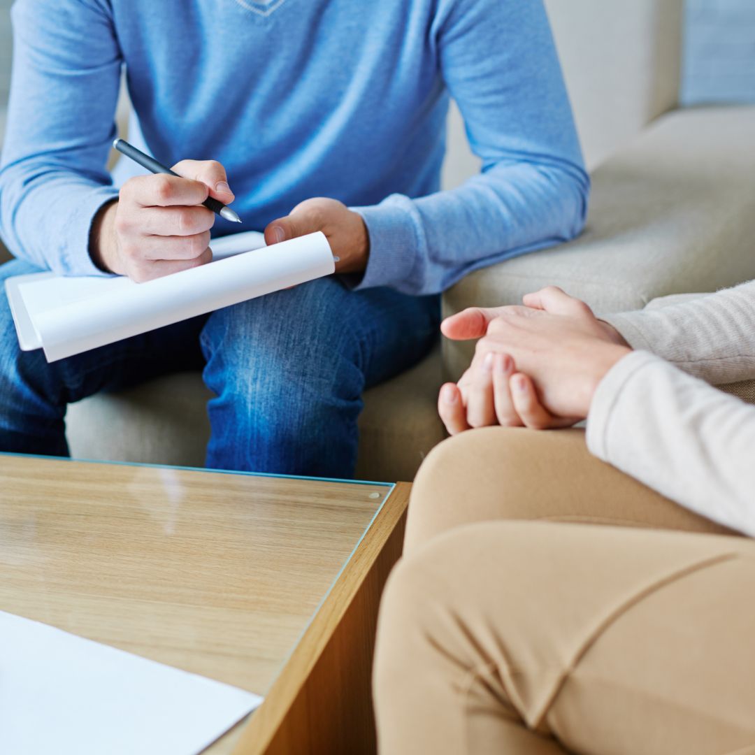 Two people sat at a table having a conversation, one is holding a cup of coffee and there is a notepad on the table.