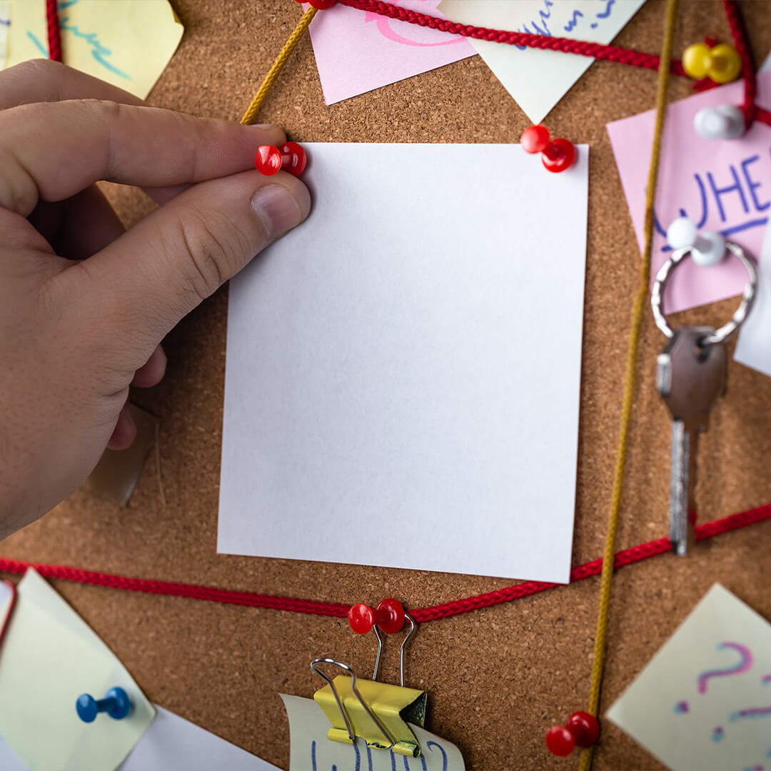 A hand, pinning a blank white paper sheet to a noticeboard.