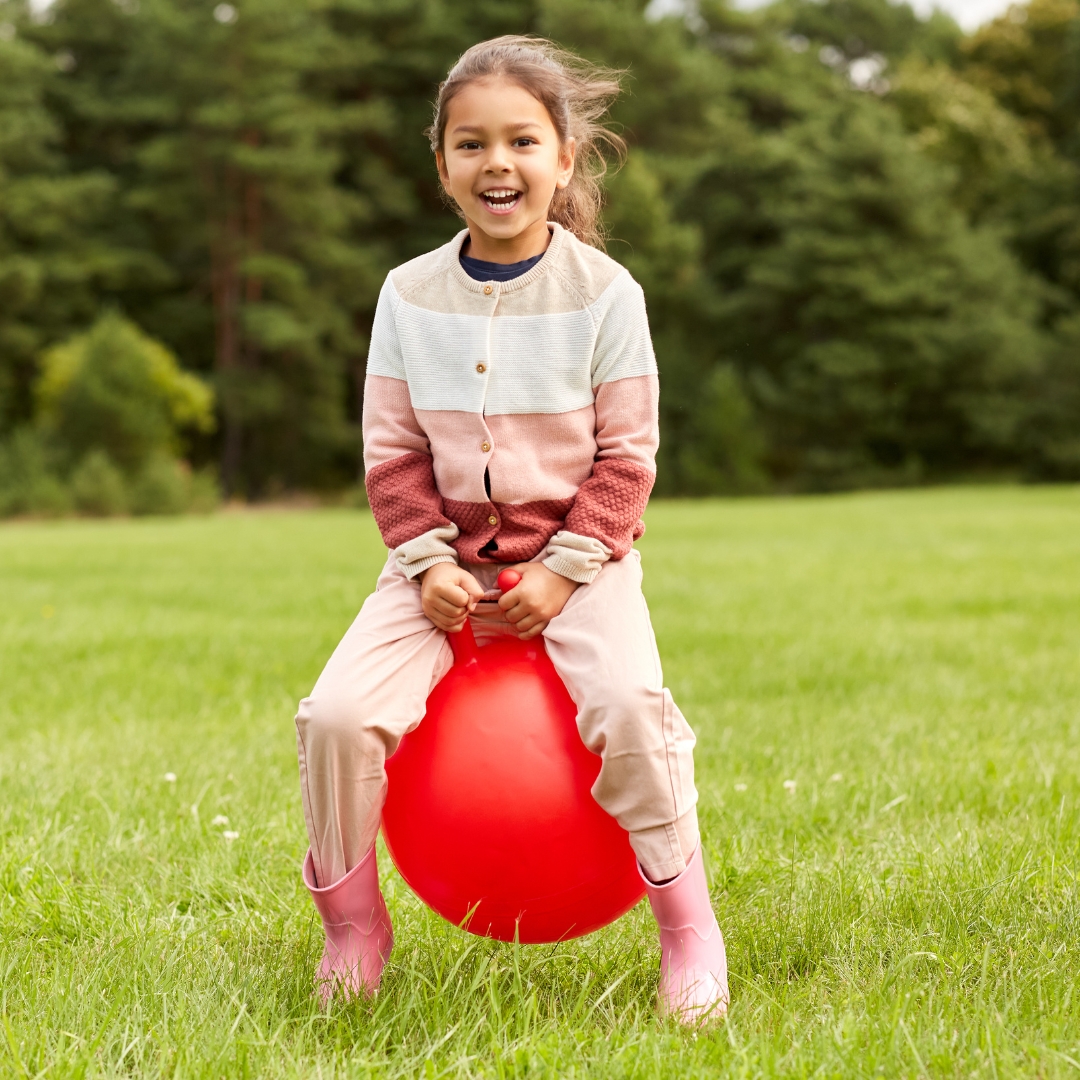 A small young girl on a bouncy hopper with two nursery nurses and four other children playing behind her.