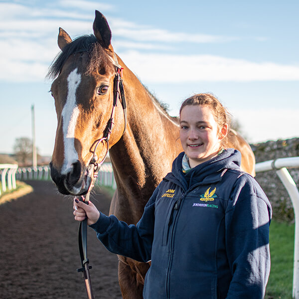 Young woman smiling whilst standing next to a brown and white horse on a race track.