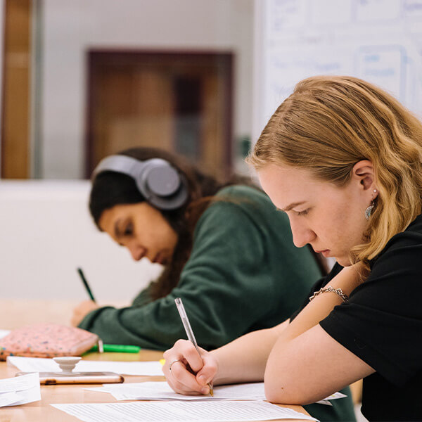 Two young women working at a desk, with a pen in their hand and their head down looking at worksheets.