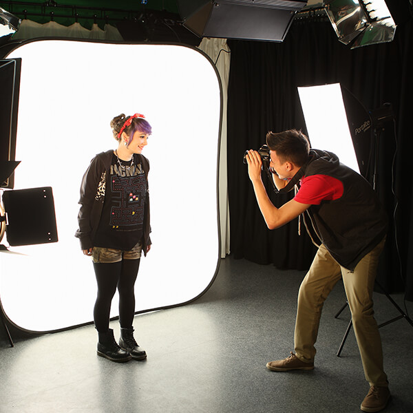 A young man taking photographs of a young woman standing in front of a light screen.