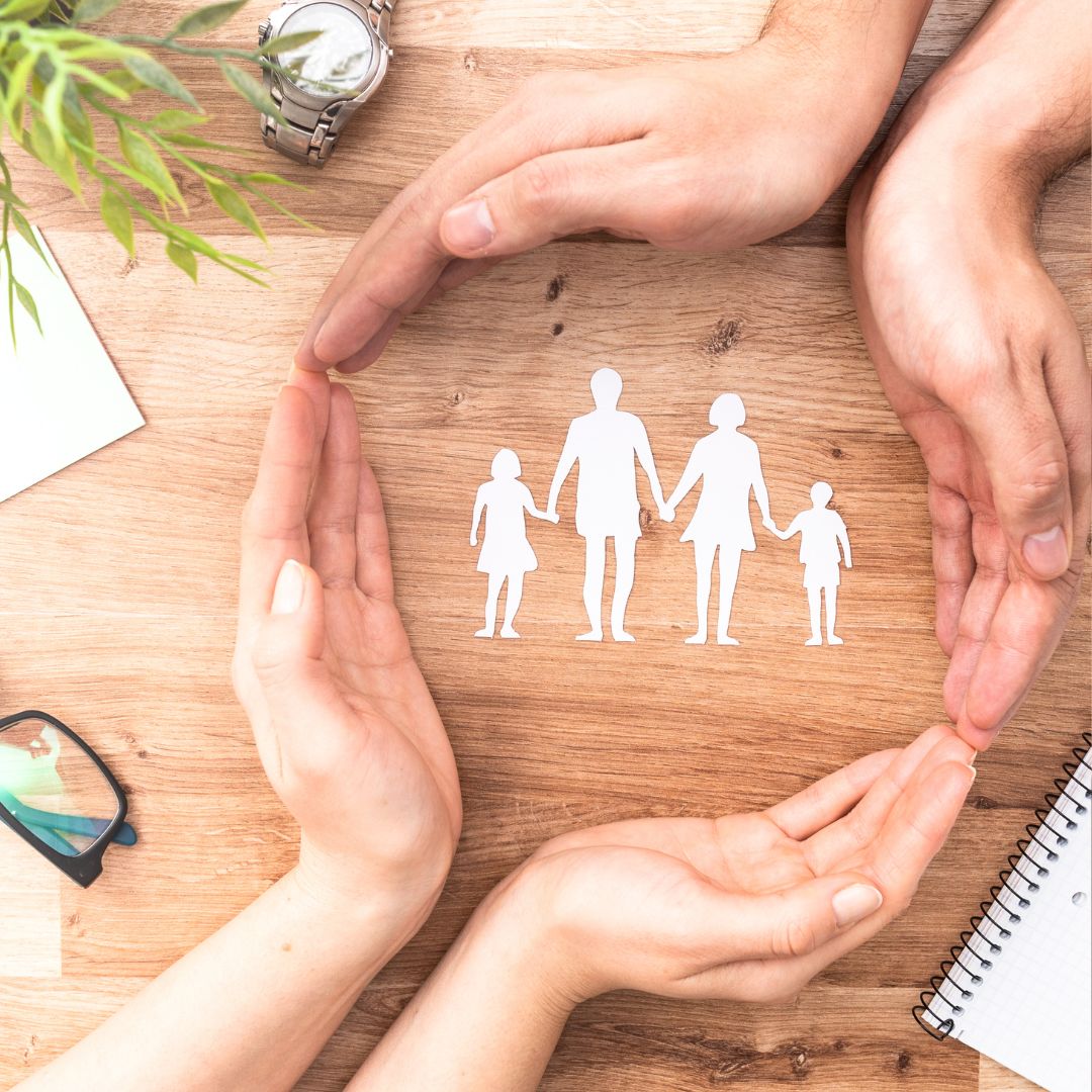 Hands surrounding cutouts of a family with two children and male and female parents holding hands