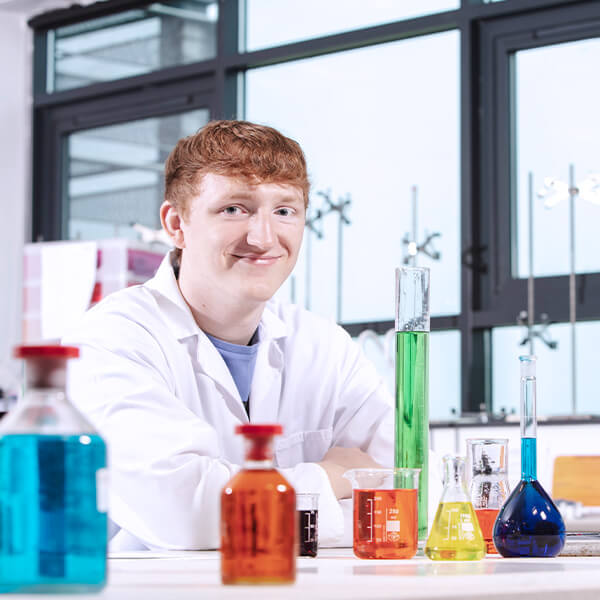 A young man wearing a white lab coat, sat at a desk that has various beakers of coloured liquid in them.