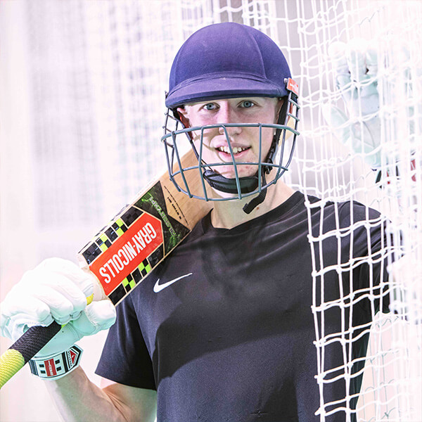 A young man wearing cricket gear and holding a cricket club, smiling.