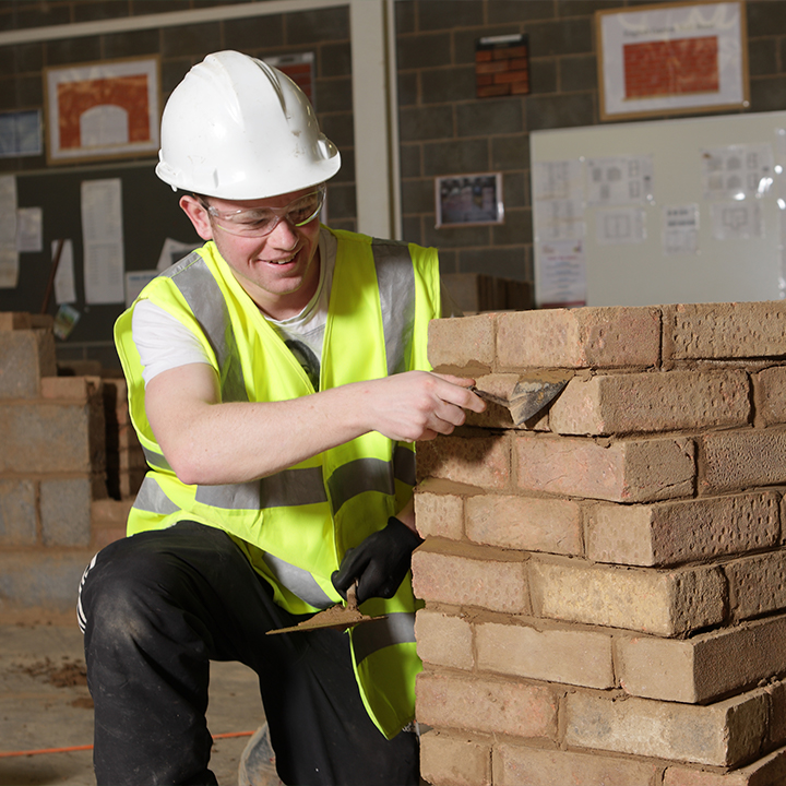 Smiling bricklaying student in high vis and hard hat using trowel to cement bricks