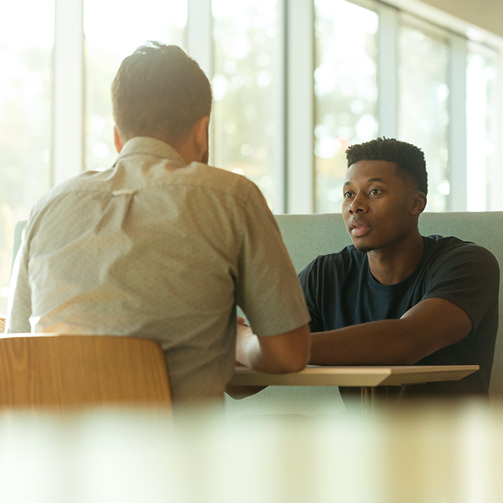 Two men talk, while sat at a table