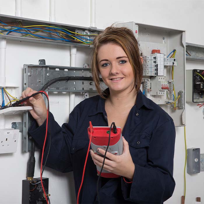 Electrical student in a blue boiler suit standing with PAT tester in front of circuitry