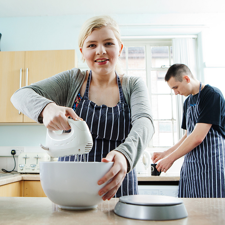 Blonde woman in apron mixes in a bowl, while man in apron behind stirs tea