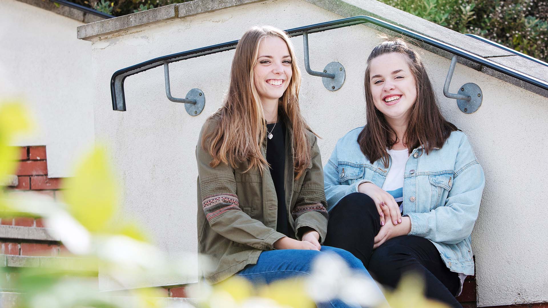 Two female students, smiling, sat outside on some steps.