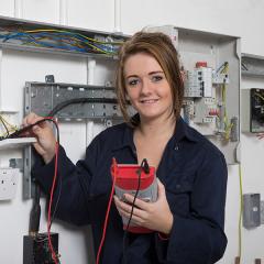 Student smiling at the camera, wearing blue overalls and holding electrical testing equipment