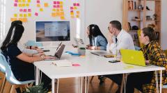 A group of five people in an office meeting with laptops and looking at a large wall mounted screen