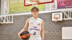 A young person holding a basketball under his right arm in a sports hall