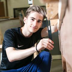 Male student holding tool smiles at the camera next to woodworking bench