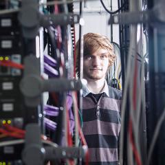 A man in a stripy shirt stands in between server equipment