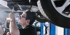 A man wearing glasses, under a raised vehicle, using a wrench