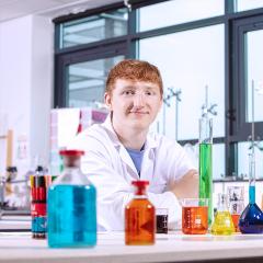 A young man in lab coat in front of chemistry equipment smiles at camera