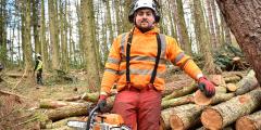 Man stood in the woods, next to a pile of cut logs, holding a chainsaw