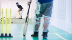 Someone in the foreground stood next to cricket stumps and someone in the background about to hit a cricket ball