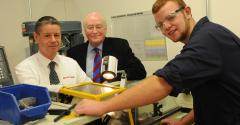 A young man wearing safety glasses next to engineering equipment with two men in the background