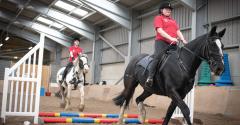 Two women in red shirts trotting on the back of a black and white horse and a black horse following the other