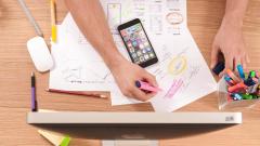 Overhead photo of a hand holding a pink highlighter with a book and phone on a desk