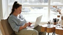 A lady sat typing with a laptop in her lap and a table in front of her with a glass of water