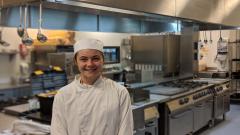 A student in chef whites, stood in a commercial kitchen, smiling at the camera