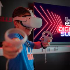 A photo of a young man wearing a VR head set with his arms out wide holding some controls.