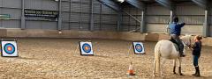 A photo of a person sat on a white horse, in an indoor equine arena, taking aim at one of three archery targets