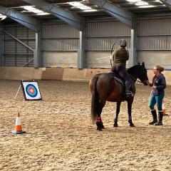 A photo of a person sat on a brown horse, in an indoor equine arena, taking aim at an archery target