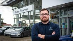 A photo of Jason Walpole wearing a blue jumper, stood with his arms folded outside Pulman Volkswagen Durham's showroom with the front of the showroom and some cars behind him.