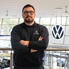 A photo of Jason Walpole wearing a blue jumper, stood with his arms folded inside Pulman Volkswagen Durham's showroom with the VW logo behind him.