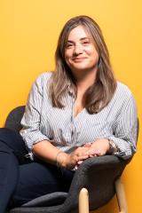 Photo of Dr Elouise Leonard-Cross sat on a black chair, looking to camera, with an orange background