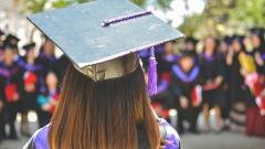 A photo of the back of someone's head, who is wearing a black university graduation mortar board hat, and looking toward a blurred crowd of people at a graduation