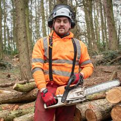 Man stood in the woods, next to a pile of cut logs, holding a chainsaw